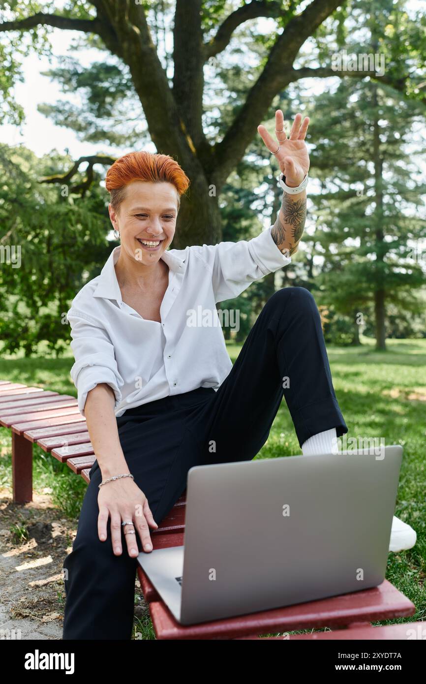 Ein Lehrer mit orangefarbenen Haaren sitzt draußen auf einer Bank, winkelt und lächelt einen Laptop an. Stockfoto