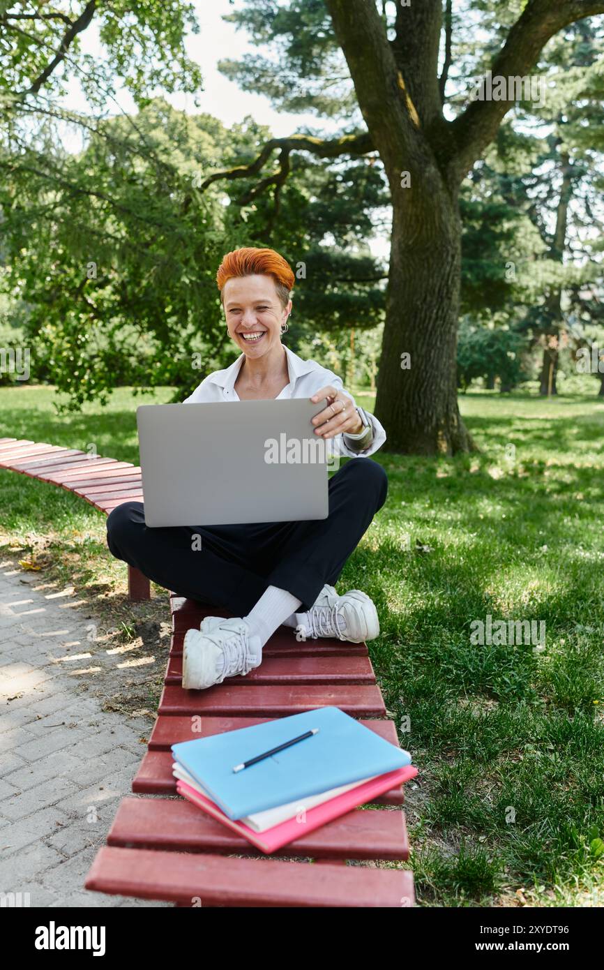 Ein Lehrer sitzt auf einer Campus-Bank mit einem Laptop, lächelt und schaut auf den Bildschirm. Stockfoto