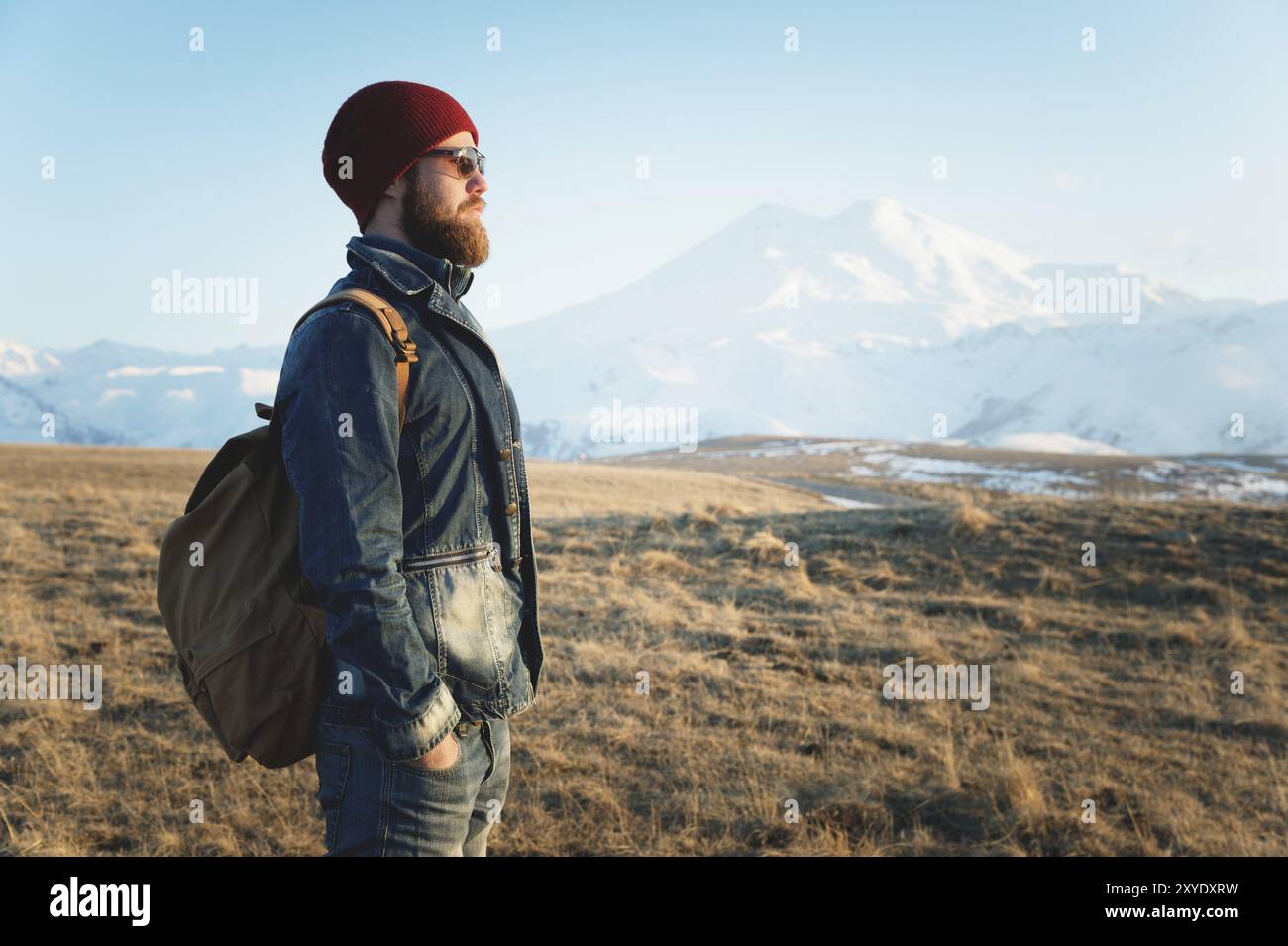 Porträt eines durchdachten, stylischen Hipsters mit Bart in Sonnenbrille und Hut mit Rucksack auf dem Hintergrund des schneebedeckten Mount Elbrus in der Stockfoto