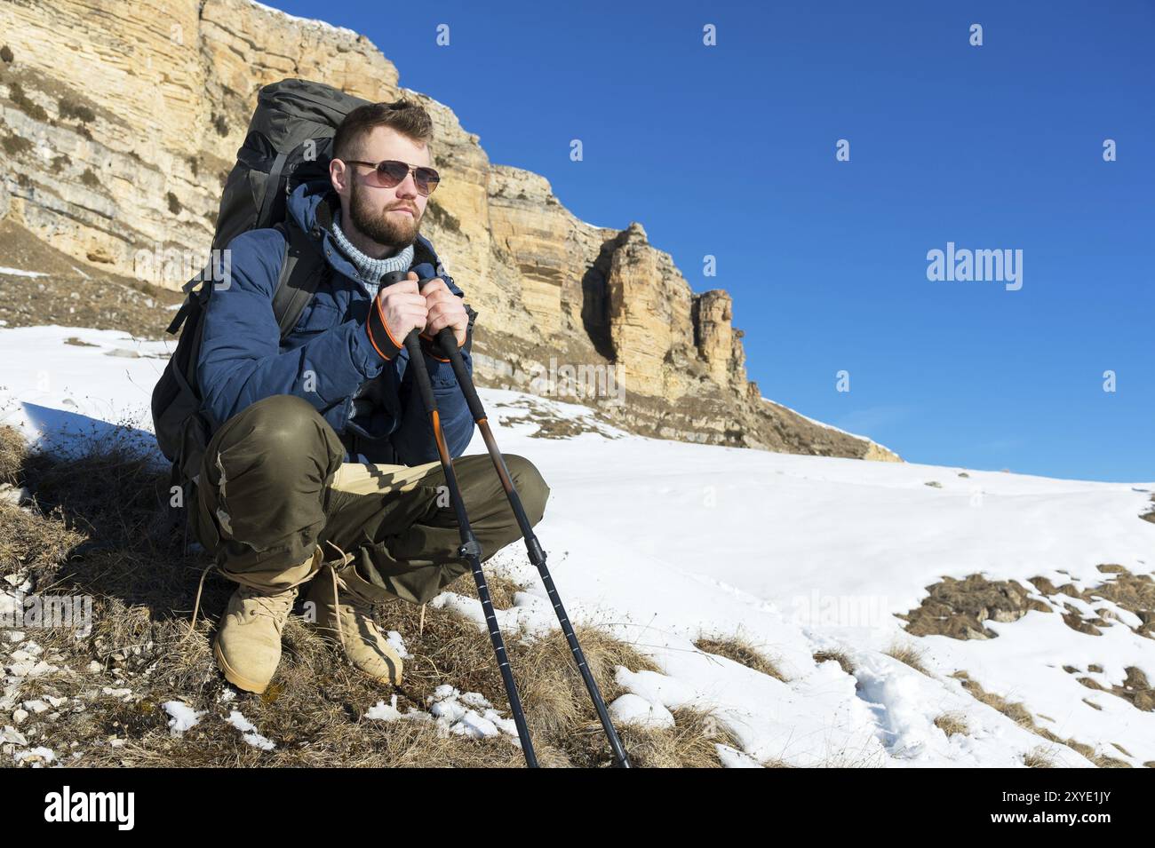 Porträt eines Hipster-Reisenden mit Bart in Sonnenbrille sitzt auf der Natur. Ein Mann wandert in den Bergen mit einem Rucksack und skandinavischem Walking sti Stockfoto