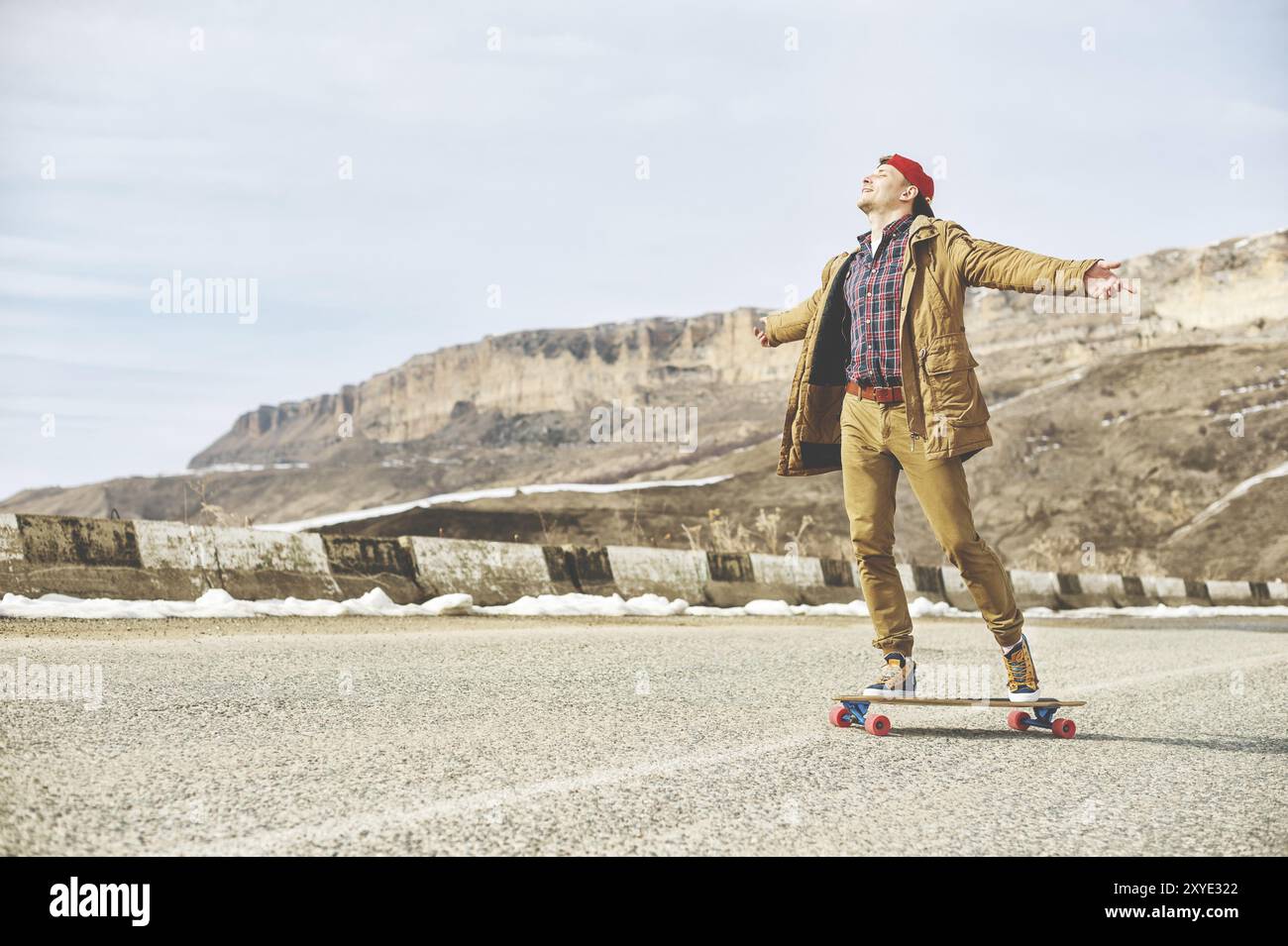 Stylischer Happy Young man in einer Mütze und Hose Jogginghose, die auf einem Longboard eine Bergstraße hinunterrollt und das Leben genießt Stockfoto