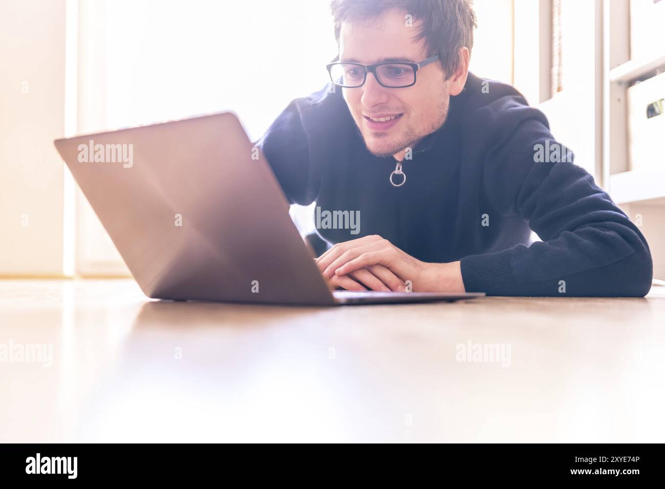 Glückliche junge Mann mit einem Laptop arbeitet auf dem Holzboden, Licht aus dem Fenster kommt Stockfoto
