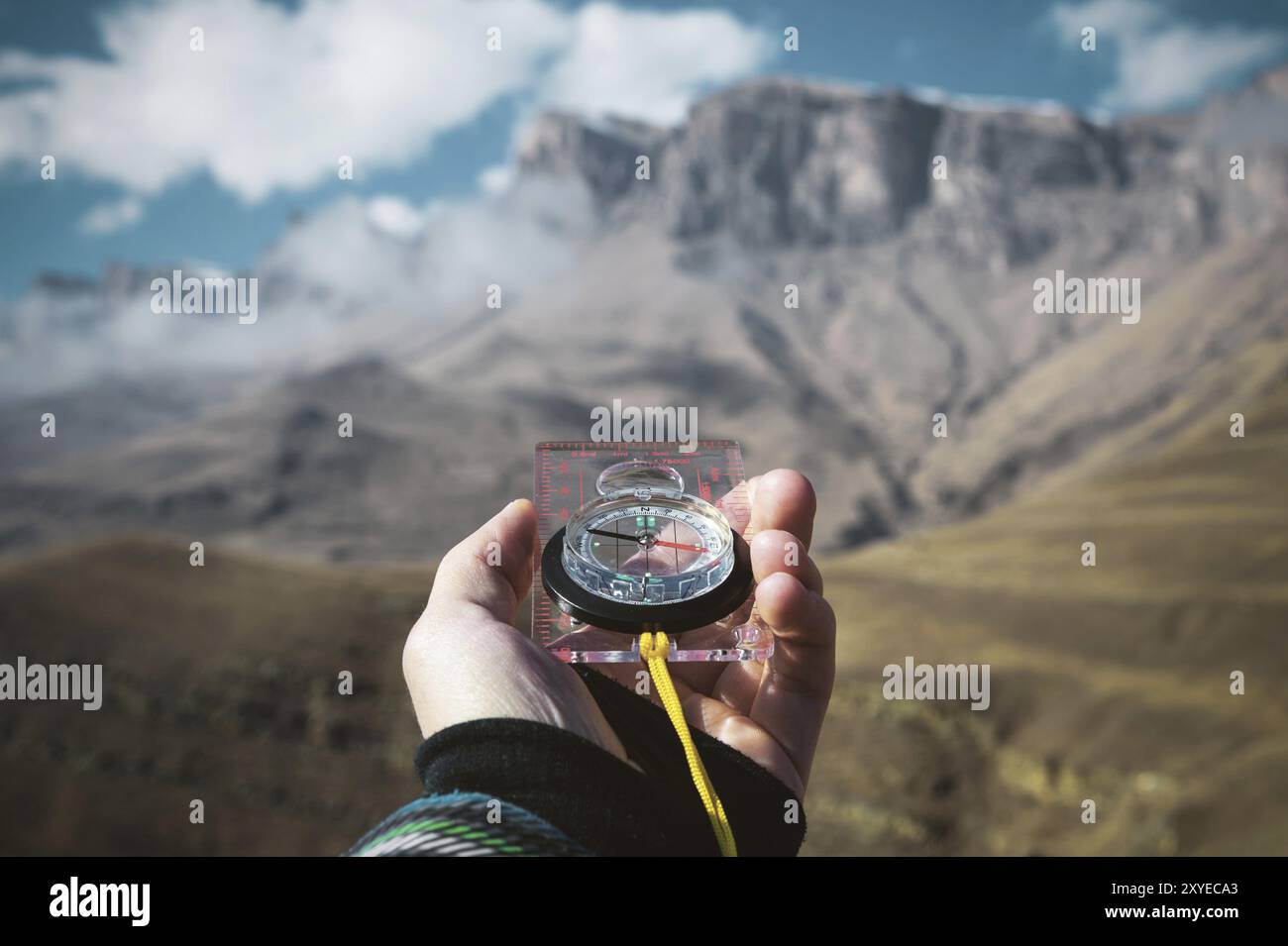 Blickpunktaufnahme. Die Hand eines Mannes aus erster Hand hält einen Kompass vor dem Hintergrund einer epischen Landschaft mit Klippen, Hügeln und einem blauen Himmel mit Stockfoto