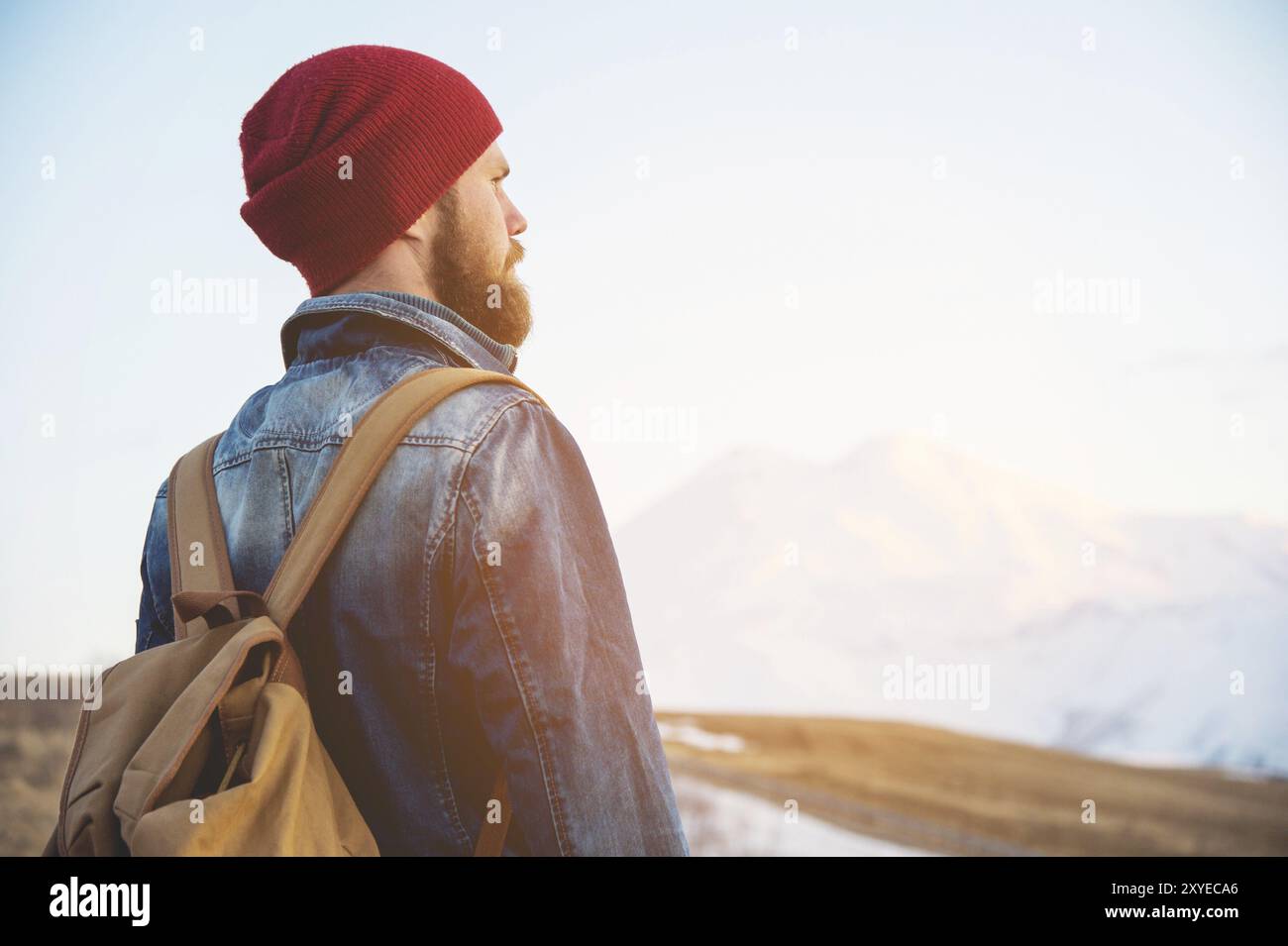 Bärtiger Touristen-Hipster-Mann mit Hut und Rucksack stehen Sie zurück auf einer Straße und beobachten Sie den Sonnenuntergang vor dem Hintergrund einer schneebedeckten mou Stockfoto