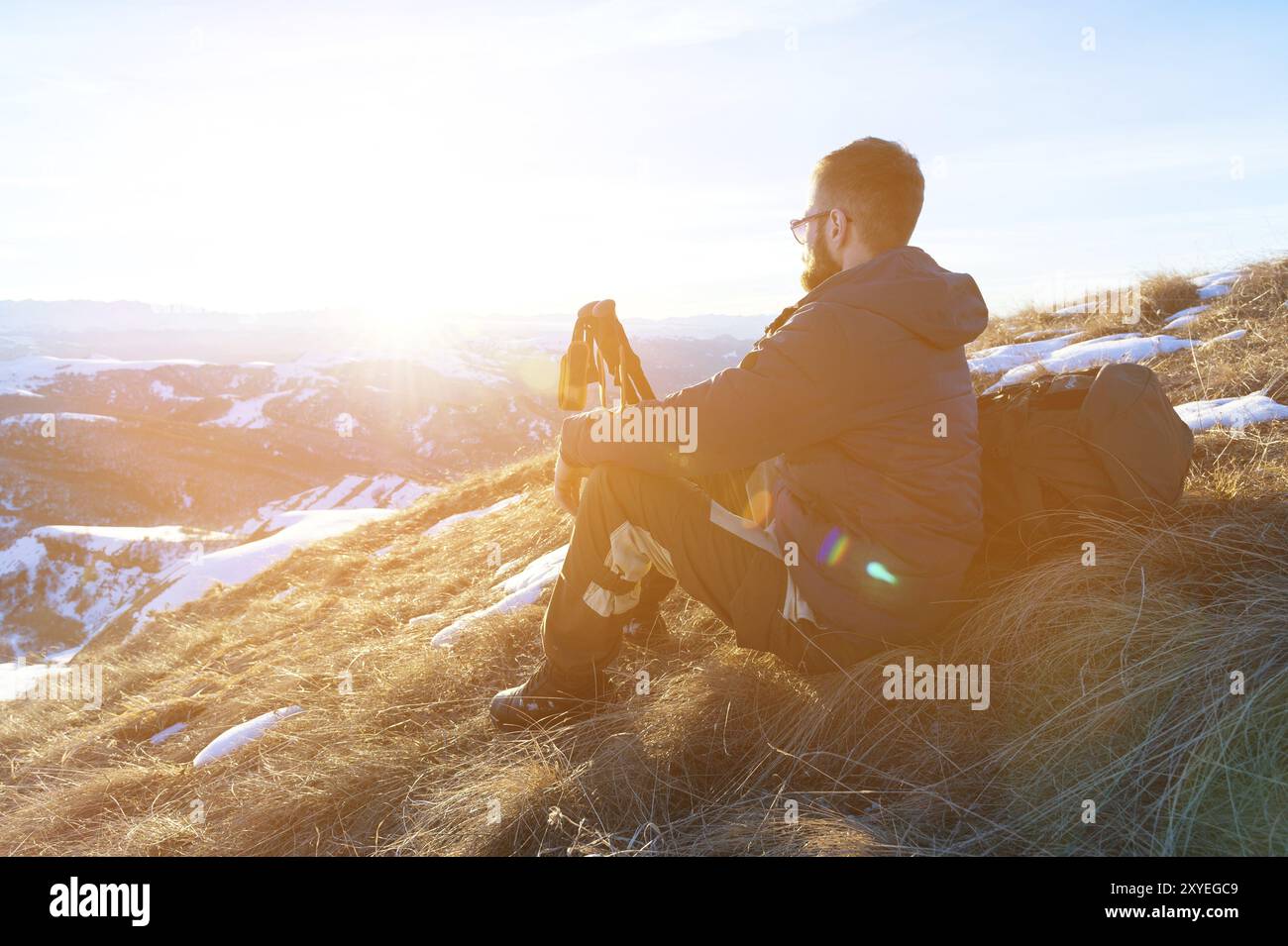 Hipster-Reisende mit Bart und Sonnenbrille sitzen im Winter auf einem gelben Gras auf einer Schieflage vor einem Hintergrund von schneebedeckten dales und einer bergigen CA Stockfoto