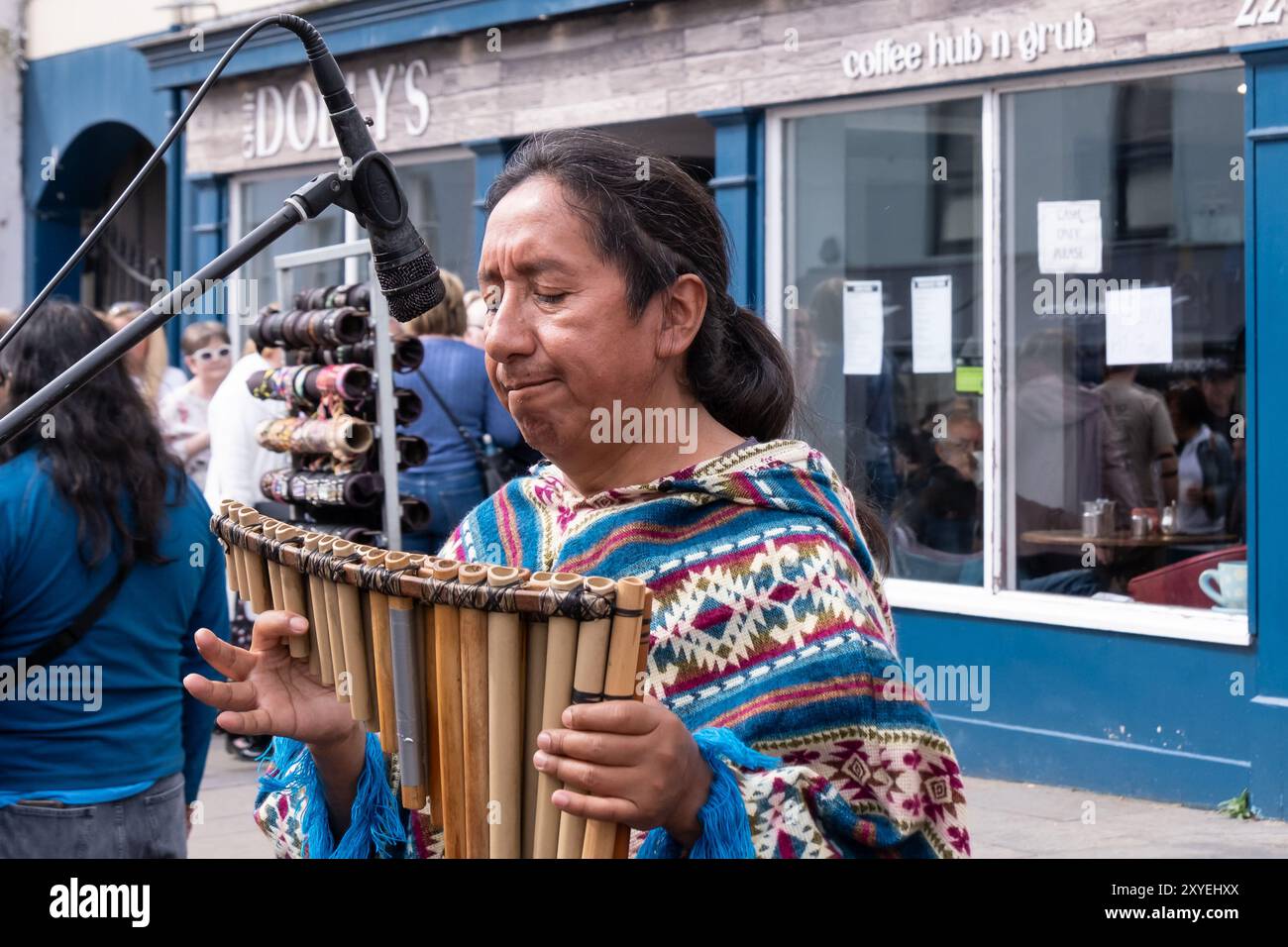 Mann mit langen Haaren und farbenfroher gemusterter Oberseite, der Pfannenpfeifen hält, traditionelle südamerikanische Flötenmusik. Ballycastle, Großbritannien - 26. August 2024. Stockfoto