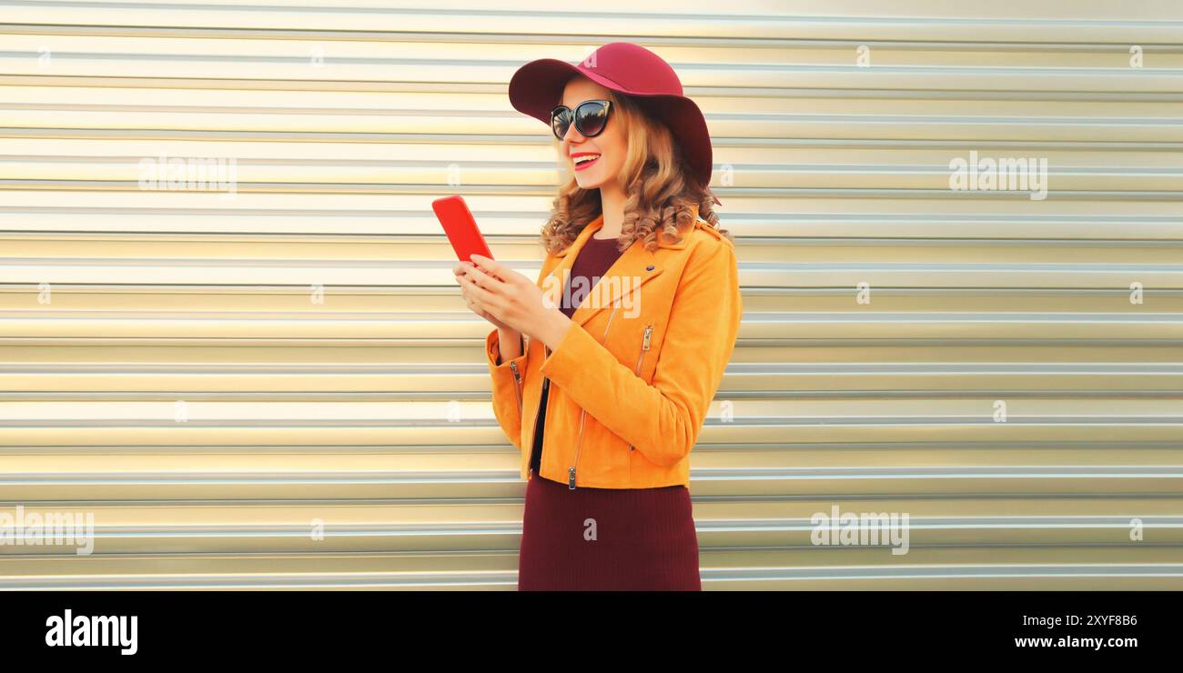 Stilvolle elegante glückliche junge Frau mit Handy, die orange Jacke, Hut und Sonnenbrille auf grauem Hintergrund trägt Stockfoto