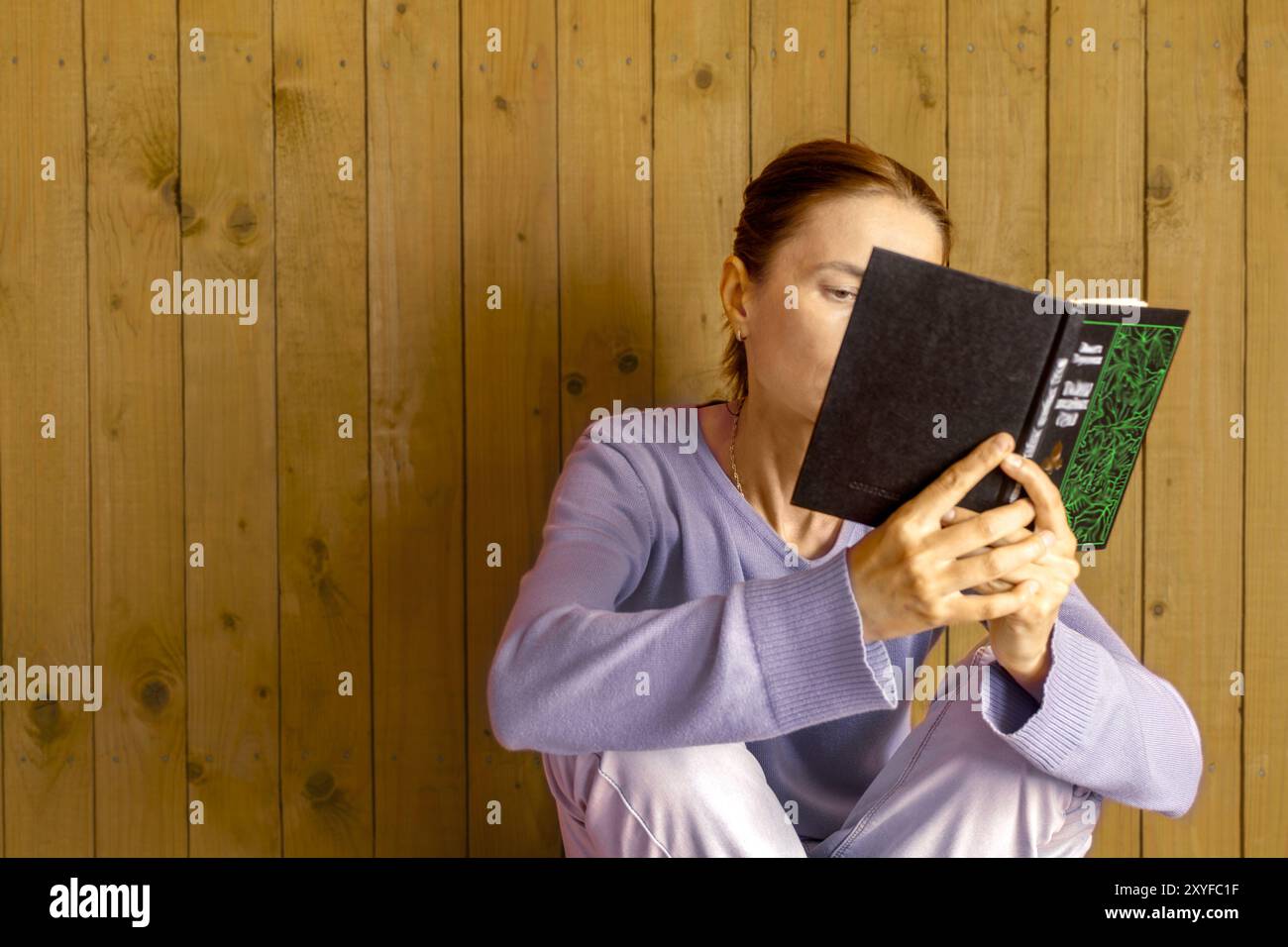 Frau in blauem Pullover, die Buch liest, sitzt an einer Holzwand im Haus. Nahaufnahme. Kopierbereich. Stockfoto