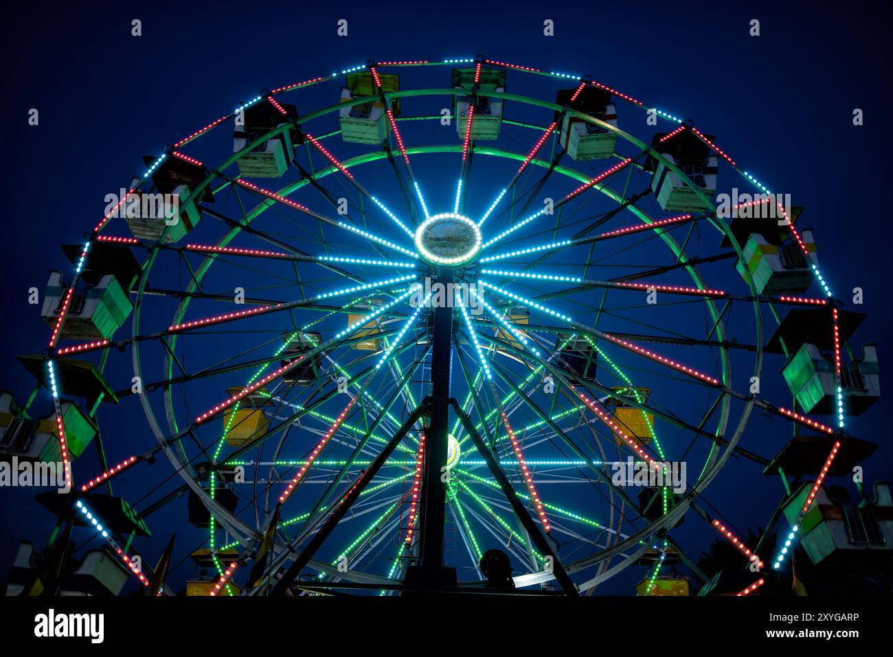 Riesenrad Arlington County Fair Arlington Virginia | ARLINGTON, Virginia — Ein lebhaft beleuchtetes Riesenrad leuchtet vor dem Abendhimmel auf der Arlington County Fair und zieht Besucher auf den Jahrmarkt im Thomas Jefferson Community Center und Park. Das kostenlose jährliche Ereignis, das seit 1977 jeden August stattfindet, entstand aus einem „Erntedankfest“, das von Gemeinschaftsgärtnern und 4-H-Mitgliedern organisiert wurde — ein bürgerlicher Ursprung, der das Wettbewerbsausstellungsprogramm der Messe bis heute prägt. Der Arlington County Fair Board, der 1976 gegründet wurde, leitete das Ereignis fast ein halbes Jahrhundert lang als eine ehrenamtliche gemeinnützige Organisation bef Stockfoto