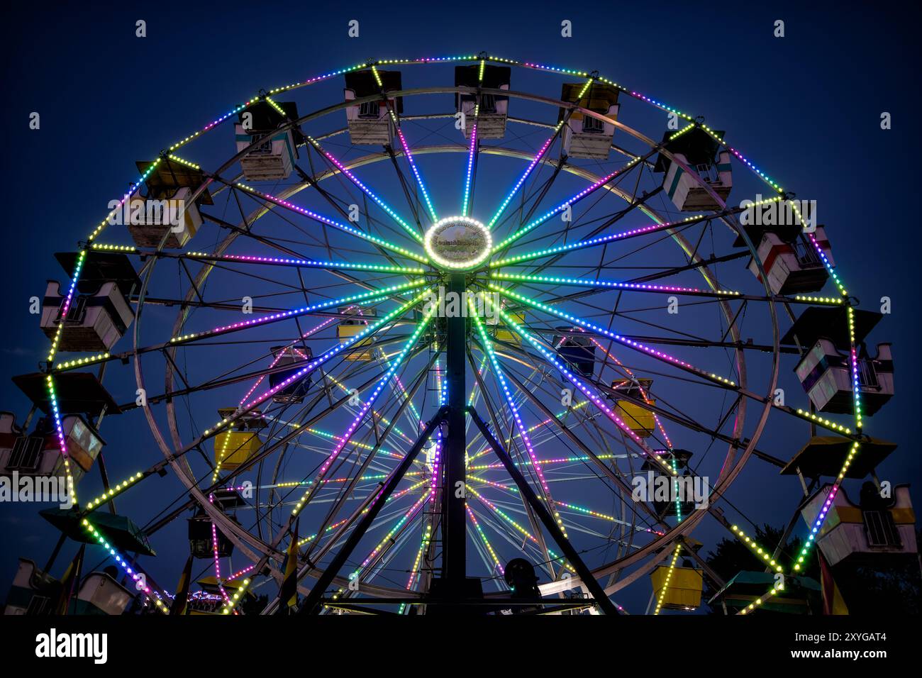 Riesenrad Arlington County Fair Arlington Virginia | ARLINGTON, Virginia — Ein lebhaft beleuchtetes Riesenrad hebt sich gegen den Abendhimmel auf der Arlington County Fair ab. Die bunten Lichter des Rades zaubern eine zauberhafte Darstellung und fangen die festliche Atmosphäre ein, wenn der Tag in die Nacht übergeht, bei dem jährlichen fünf Tage dauernden Ereignis, das jeden August im Thomas Jefferson Community Center und Park stattfindet. Die Messe, die 1977 begann und kostenlos besucht werden kann, wird von den Veranstaltern als eines der größten kostenlosen Ereignisse an der Ostküste beschrieben. Im Jahr 2025 zeigte die Messe zwei Riesenräder neben ihren Fahrgeschäften auf dem Festplatz, Wett Stockfoto