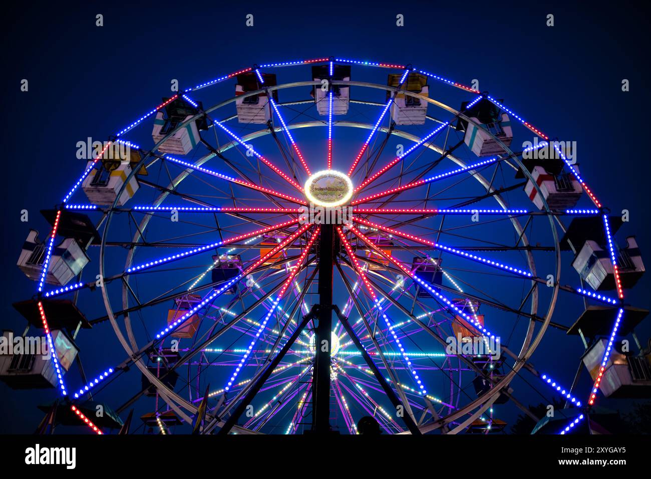 Arlington Virginia // ARLINGTON, Virginia — Ein leuchtend beleuchtetes Riesenrad hebt sich auf der Arlington County Fair vor dem Abendhimmel hervor. Die farbenfrohen Lichter des Rades sorgen für eine blendende Präsentation, die die festliche Atmosphäre der Messe einfängt, wenn der Tag in die Nacht verwandelt wird. Diese legendäre Karnevalsfahrt dient als Herzstück des jährlichen Gemeindeereignisses und bietet sowohl aufregende Ausblicke für Fahrer als auch eine malerische Szene für Märchenbesucher. Stockfoto