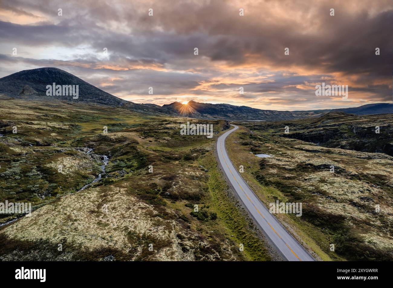 Aus der Vogelperspektive auf eine Straße, die sich durch die sanften Hügel schlängelt, den Sonnenuntergang hinter den Bergen, der einen warmen Glanz über die beschauliche nordische Landschaft wirft Stockfoto