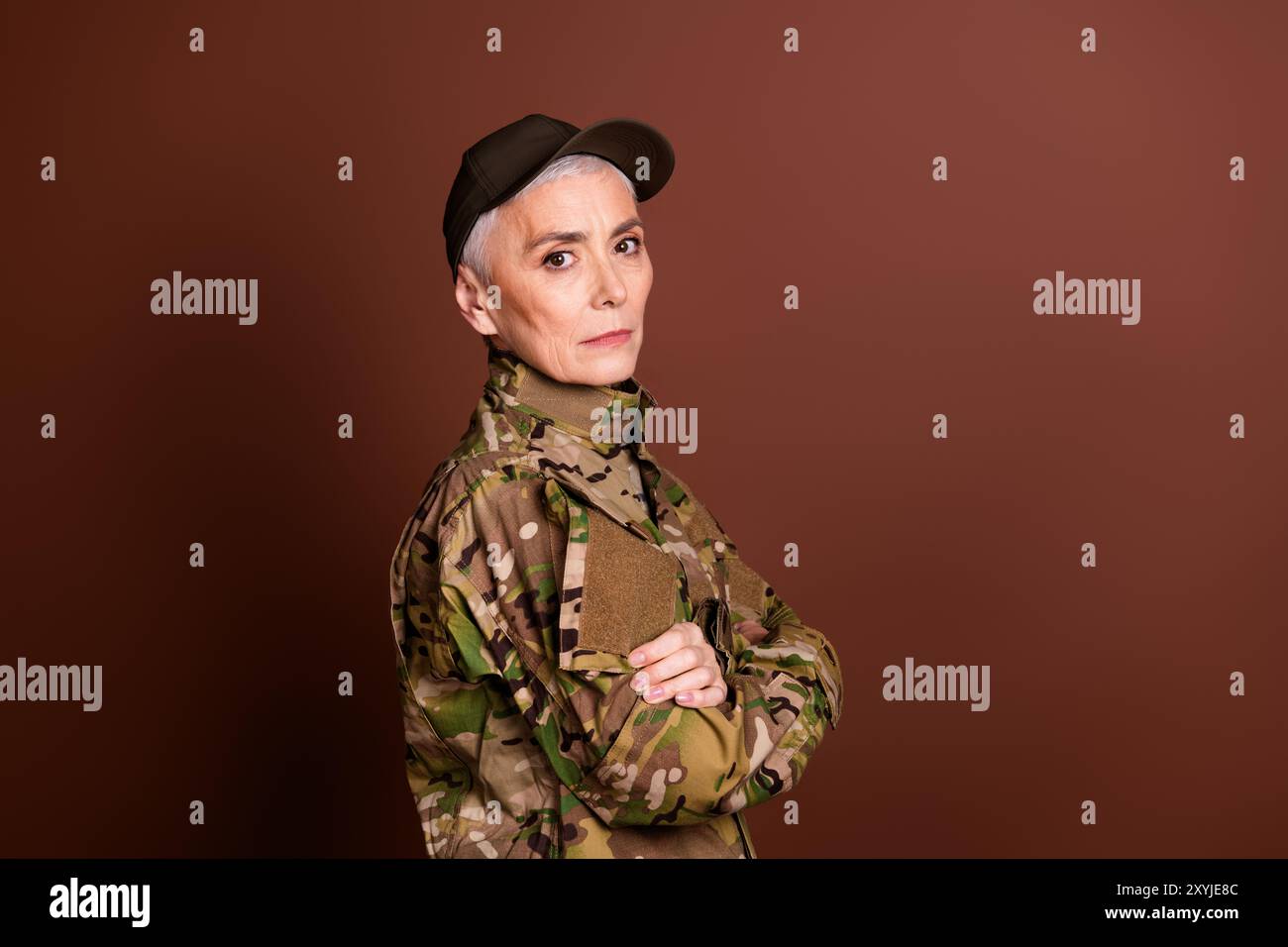 Foto einer seriösen Senior-Frau, die Tarnarme trägt, durchquert den leeren Raum, isolierter brauner Hintergrund Stockfoto