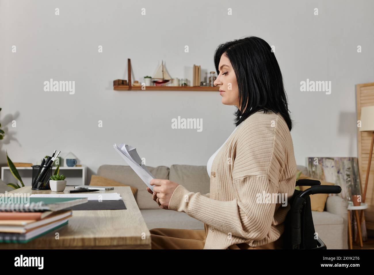 Eine Frau sitzt in einem Rollstuhl an einem Schreibtisch und liest einige Zeitungen. Stockfoto