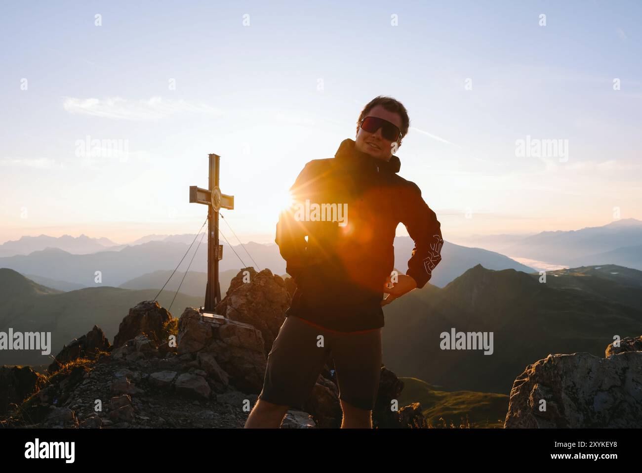 Mann auf einem Berg, Sonnenaufgang, mit einem Gipfelkreuz im Hintergrund. Stockfoto