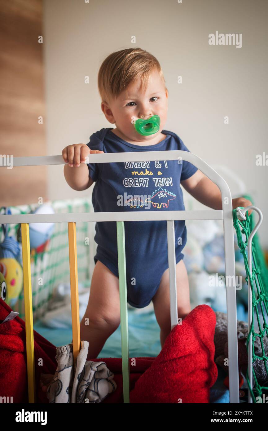 Ein fröhliches Kleinkind mit einem Schnuller erkundet sein farbenfrohes Kinderbett, das hoch in einer Vielzahl von weichen Spielzeugen und Decken steht und während eines s pure Freude ausstrahlt Stockfoto