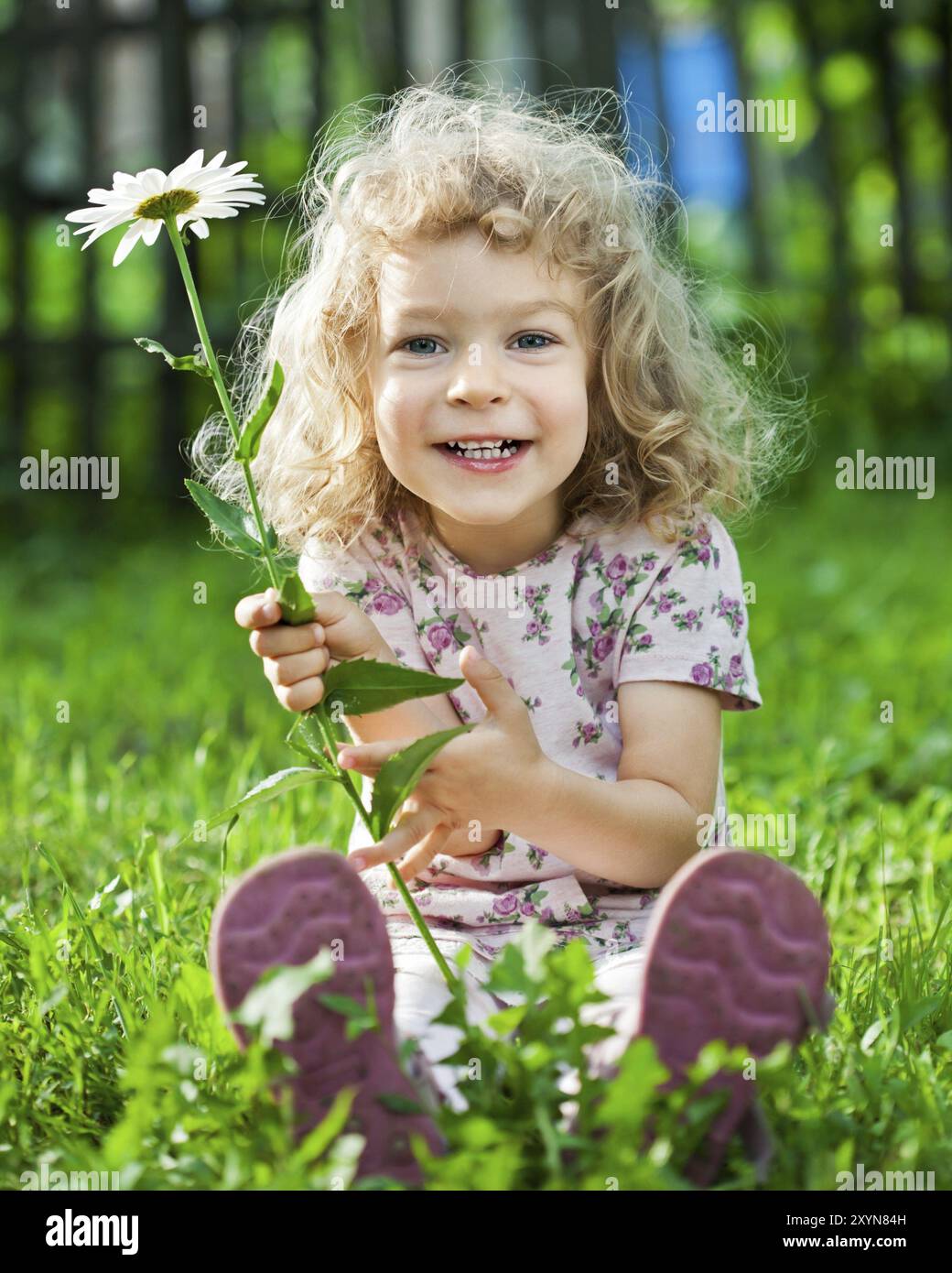 Glückliches lächelndes Kind mit Blume, das draußen im Frühlingsgarten auf grünem Gras sitzt Stockfoto