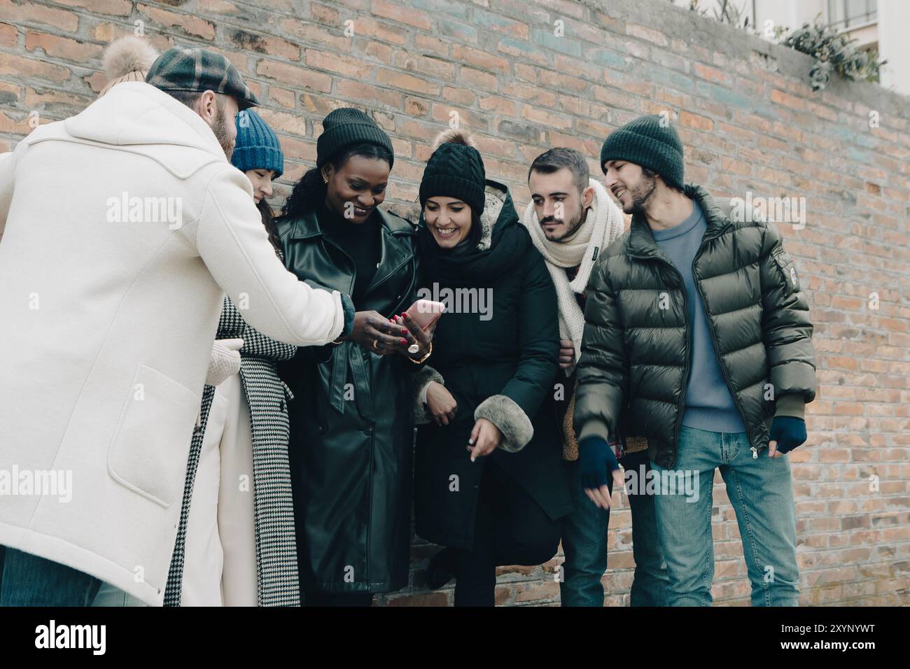 An einem kalten Wintertag draussen sitzen Freunde zusammen, schauen auf ein Smartphone und lächeln Stockfoto