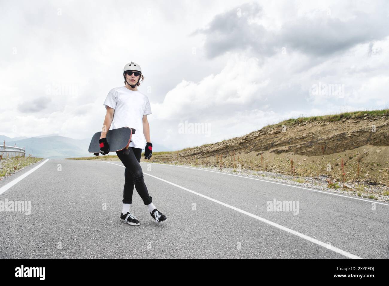 Ein junger Mann, ein Skater in einem Helm mit Brille und Handschuhen wartet auf ein Rennen auf einer Landstraße mit einem Longboard in der Hand Stockfoto