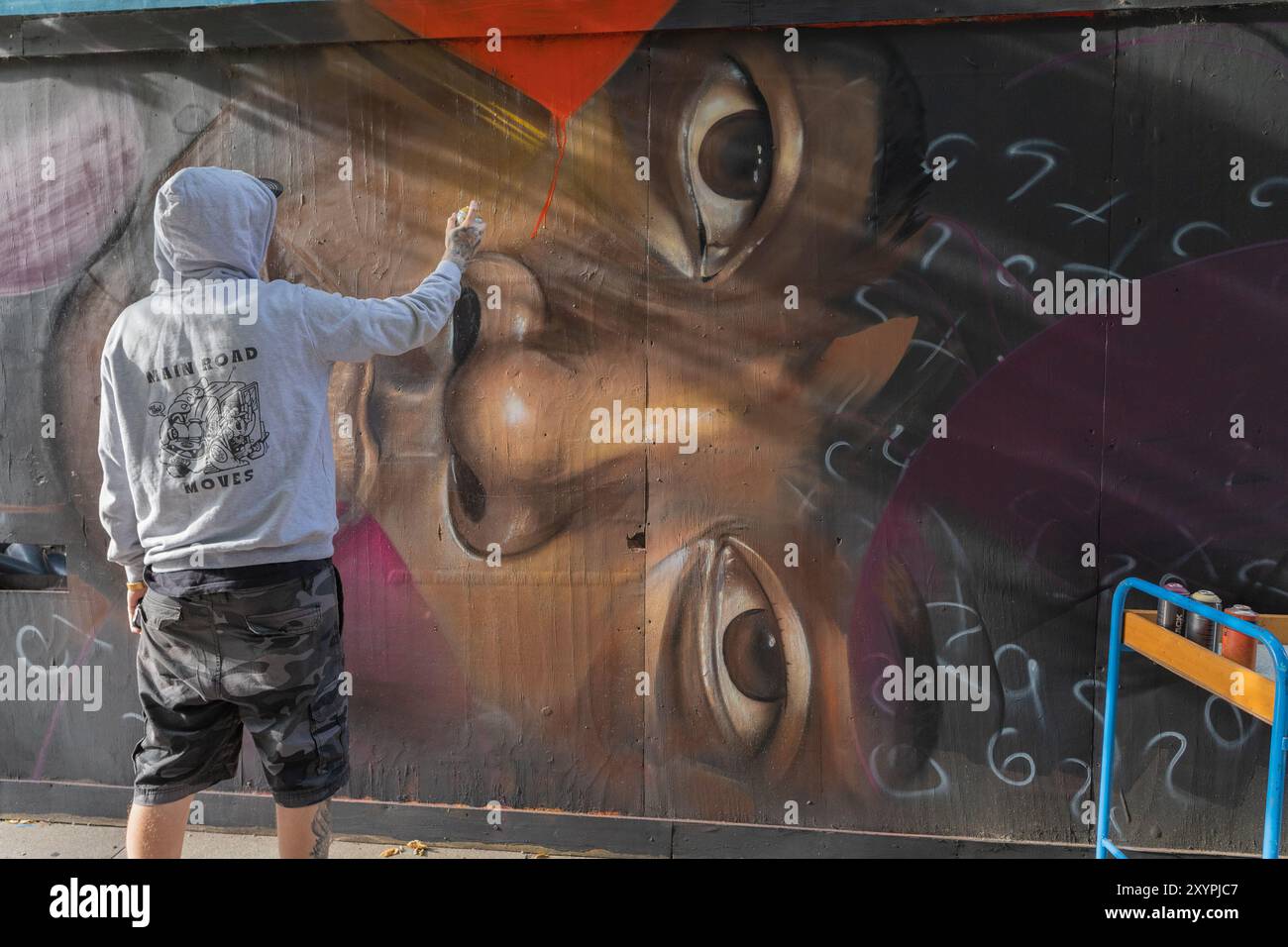 Southend on Sea, Großbritannien. 30. August 2024. Ein Wandkünstler in einem grauen Hoodie und Camouflage-Shorts besprüht ein großes, realistisches Gesicht auf einer öffentlichen Wand in Southend-on-Sea. Das Bildmaterial enthält stilisierte Elemente und ein abstraktes Skript. Eine blaue und orangefarbene Barriere schließt den Bereich ab. City Jam Day One endet mit Künstlern, die an ihrer kreativen Arbeit arbeiten. Penelope Barritt/Alamy Live News Stockfoto