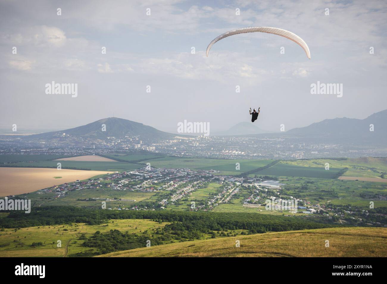 Professioneller Paragleiter im Kokon-Anzug fliegt hoch über dem Boden gegen den Himmel und Felder mit Bergen Stockfoto