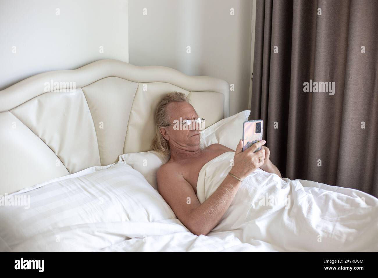 Reifer blonder Mann mit Brille liegt auf dem Bett unter der Decke und liest Nachrichten am Telefon. Erholung und Wochenenden. Stockfoto