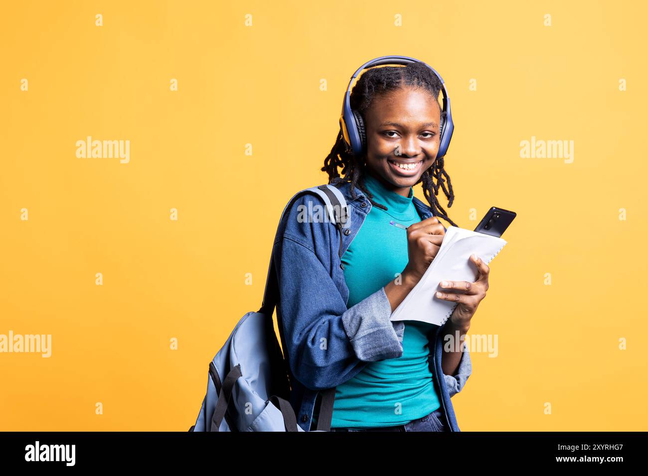 Porträt eines lächelnden Schülers, der Hausaufgaben schreibt, online über Smartphone recherchiert und Musik hört, Studiohintergrund. Jolly Girl macht Notizen für die Schulbeurteilung und genießt Lieder auf Kopfhörern Stockfoto