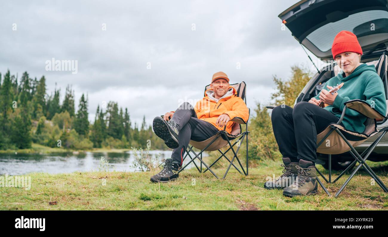 Friedliche norwegische Campingszene mit Vater und Sohn am See. Sie entspannen in Stühlen, während Sohn Ukulele spielt, umgeben von der Natur, lachend genießen Stockfoto