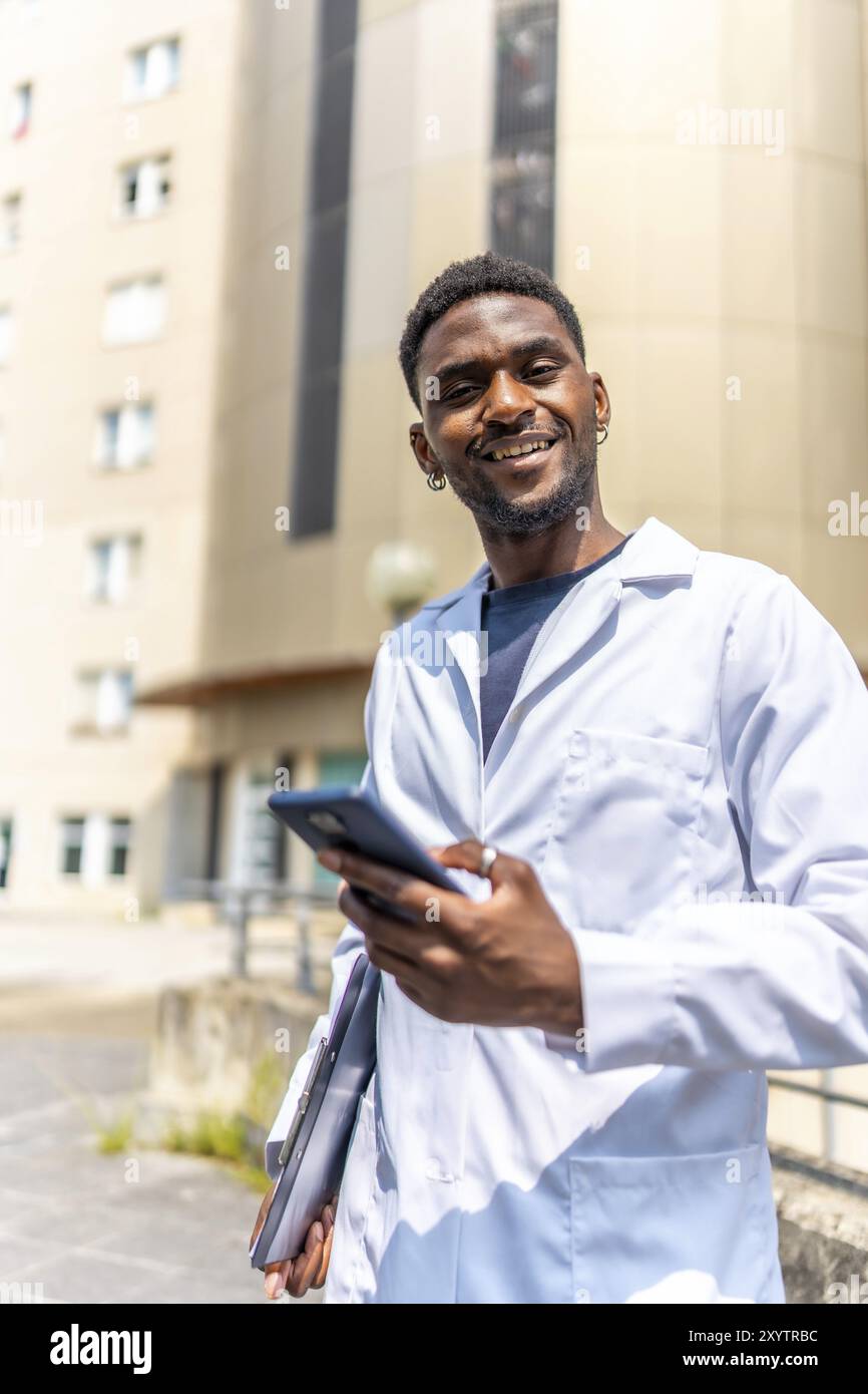 Vertikales Foto eines fröhlichen jungen afrikanischen Arztes mit Telefon, das vor dem Krankenhaus steht Stockfoto