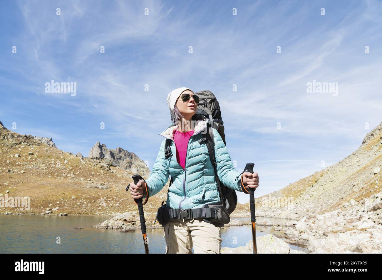 Wandermädchen in Sonnenbrille Daunenjacke und Hut mit Rucksack und Bergausrüstung mit Tracking-Stöcken in den Händen schaut auf die wunderschöne Aussicht auf eine Stockfoto