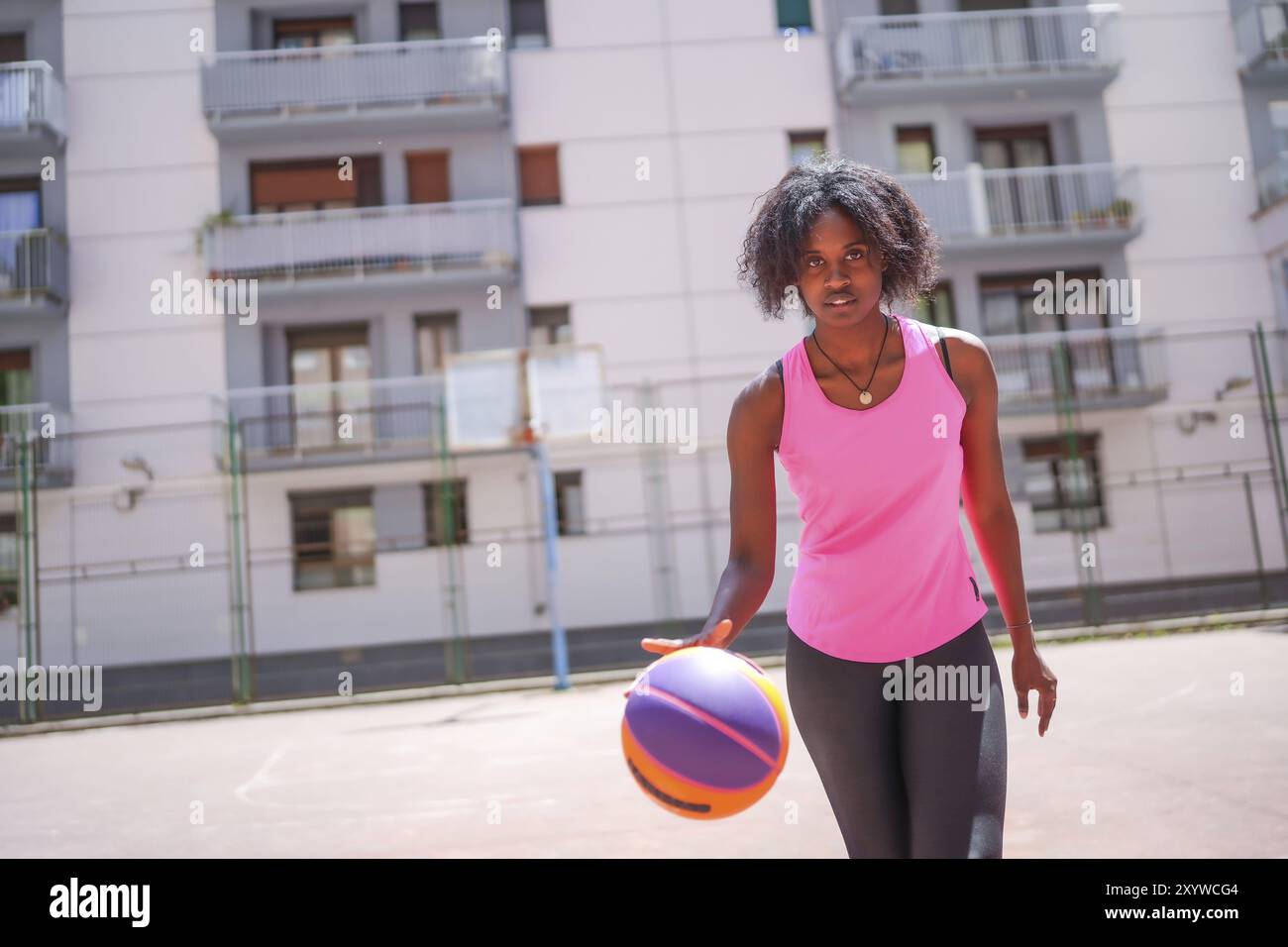 Selbstbewusste Afro-Frau, die in die Kamera blickt, während sie auf einem städtischen Basketballfeld dribbelt Stockfoto