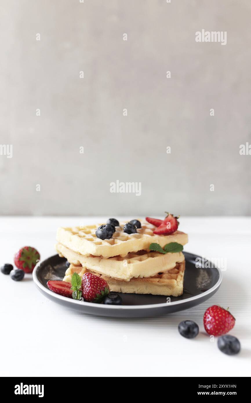 Stapel Waffeln auf einem Teller auf dem weißen Tisch mit Blaubeeren, gehackten Erdbeeren- und Minzblättern, grauem Hintergrund. Stockfoto