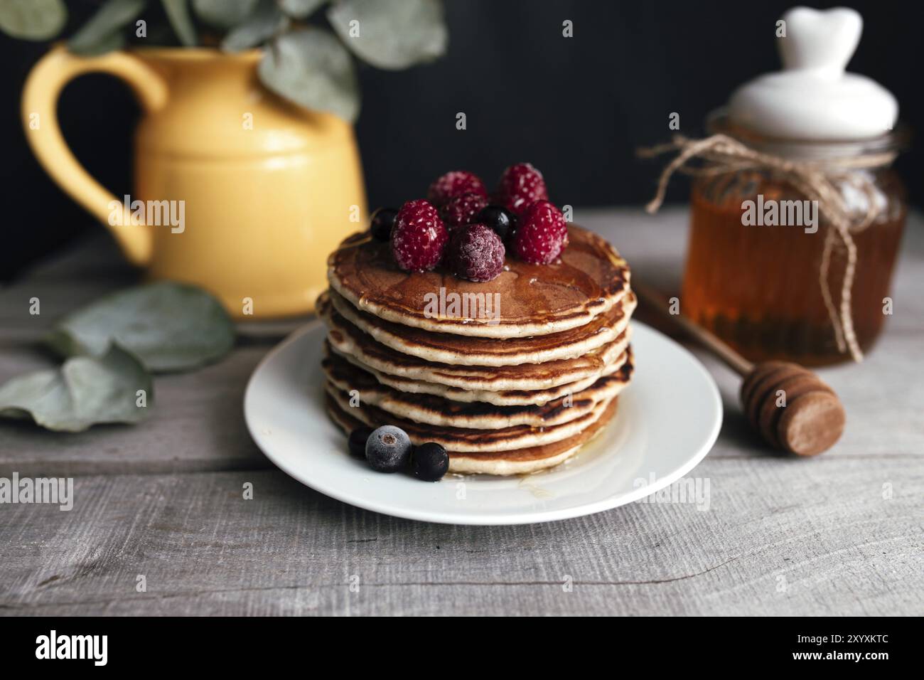 Saftige Pfannkuchen mit Beeren und Honig auf einem weißen Teller, Löffel, Glas, Holztisch, gelbe Vase mit Eukalyptus. Stockfoto