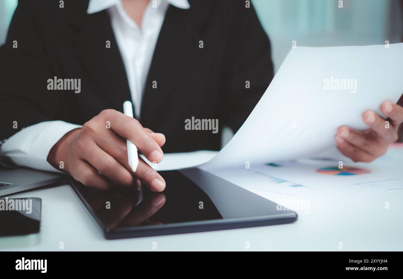 Geschäftsfrau in schwarzem Anzug, die an Laptop-Computer, Papierdokumenten, Tablet und Rechner arbeitet, im Büro mit einem dunklen Hintergrund das Tablet mit den Händen berührt, Stockfoto