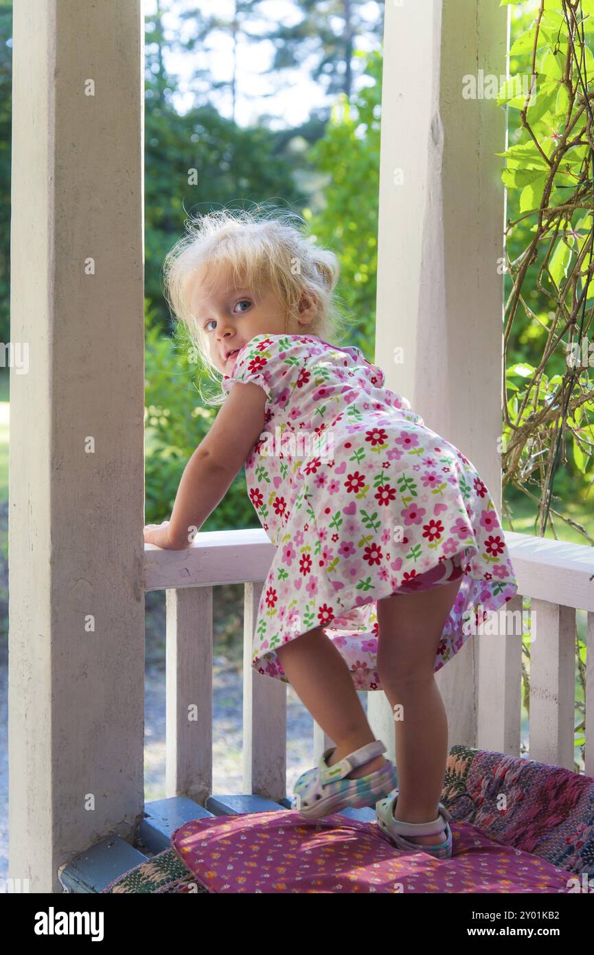 Ein süßes kleines Mädchen im Sommerkleid steht auf der Veranda. Wundervolle, sommerliche, lichtdurchflutete Atmosphäre. Süßes blondes Mädchen, das einen süßen Sommer trägt Stockfoto