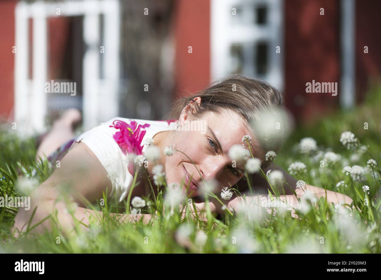 Eine junge Frau liegt auf einer Sommerwiese, lächelt und entspannt. Im Hintergrund ist ein idyllisches schwedisches Haus zu sehen. Das Sonnenlicht gibt den ganzen Co Stockfoto
