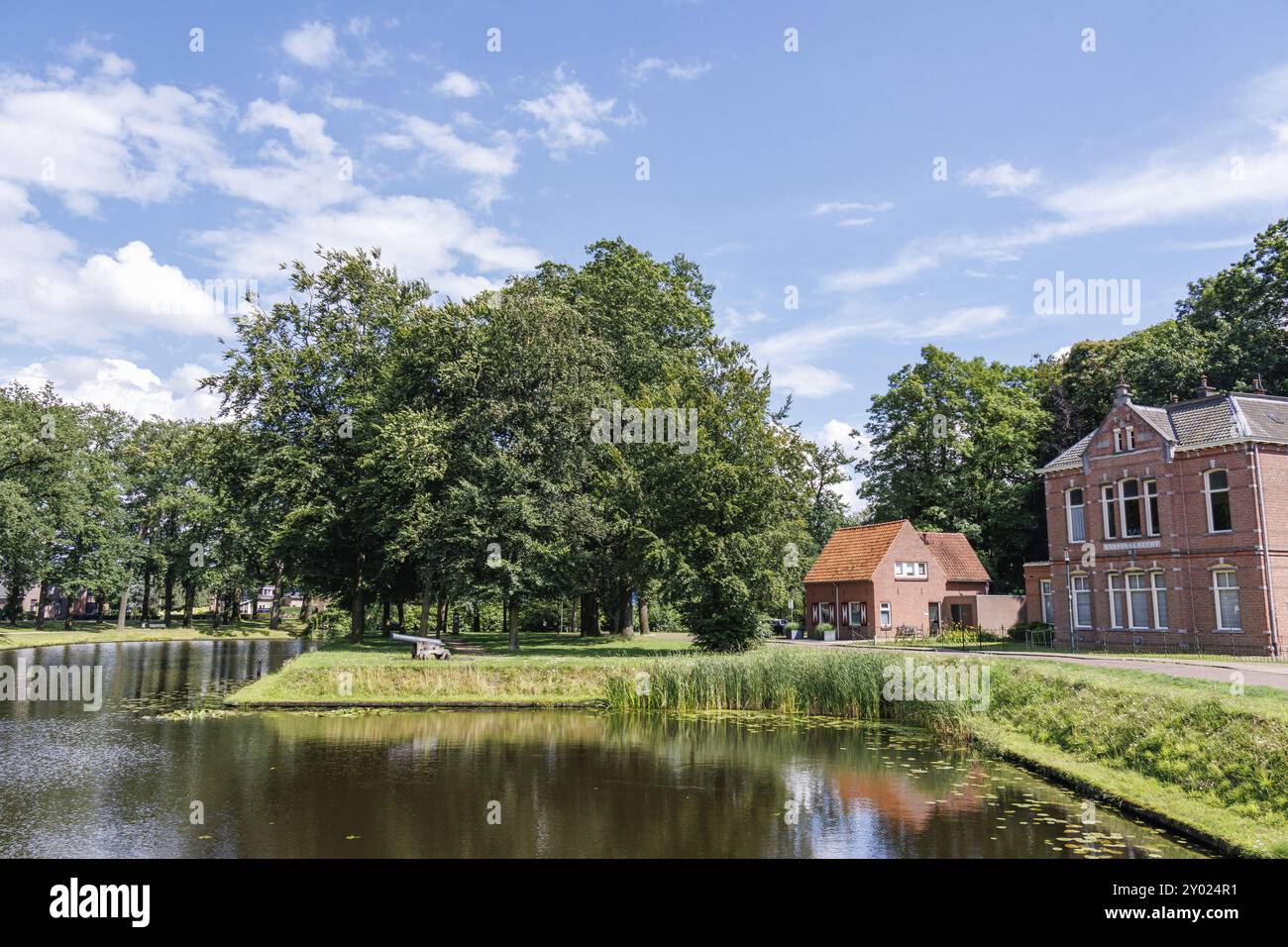 Backsteinbau und Haus auf einem Teich mit umliegendem Rasen und Bäumen unter leicht bewölktem Himmel, aalten 2 Stockfoto