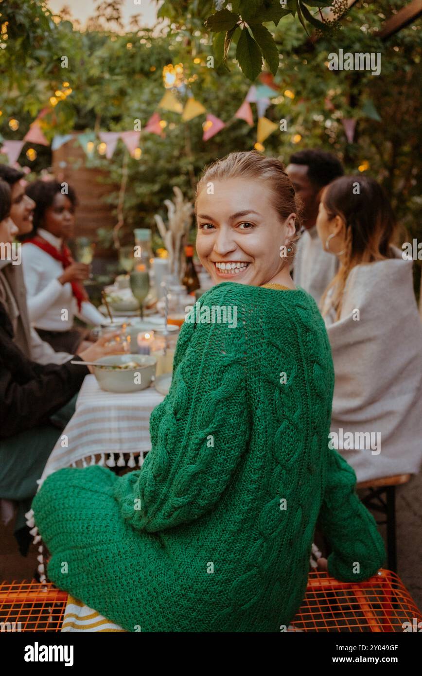 Porträt einer glücklichen Frau in grünem Pullover, der über die Schulter schaut, während er mit Freunden am Esstisch sitzt Stockfoto