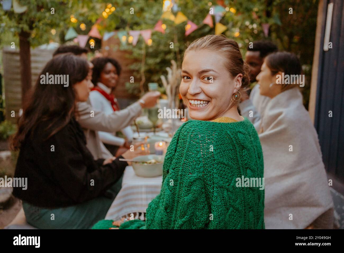 Porträt einer fröhlichen jungen Frau, die über die Schulter schaut, während sie mit Freunden im Hinterhof sitzt Stockfoto