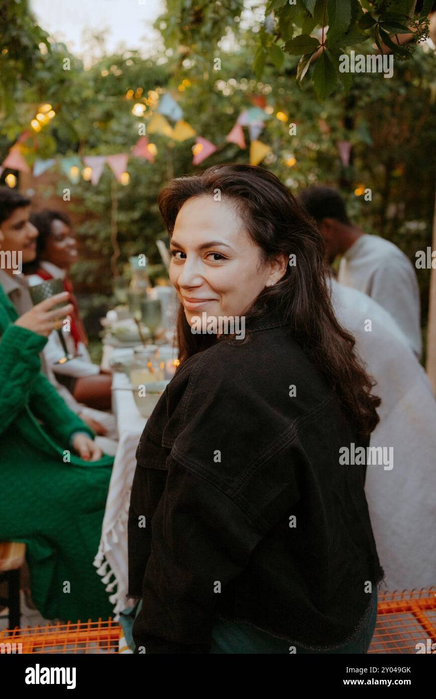 Porträt einer lächelnden Frau mit langen Haaren, die über die Schulter schaut, während sie mit Freunden am Esstisch im Hinterhof sitzt Stockfoto