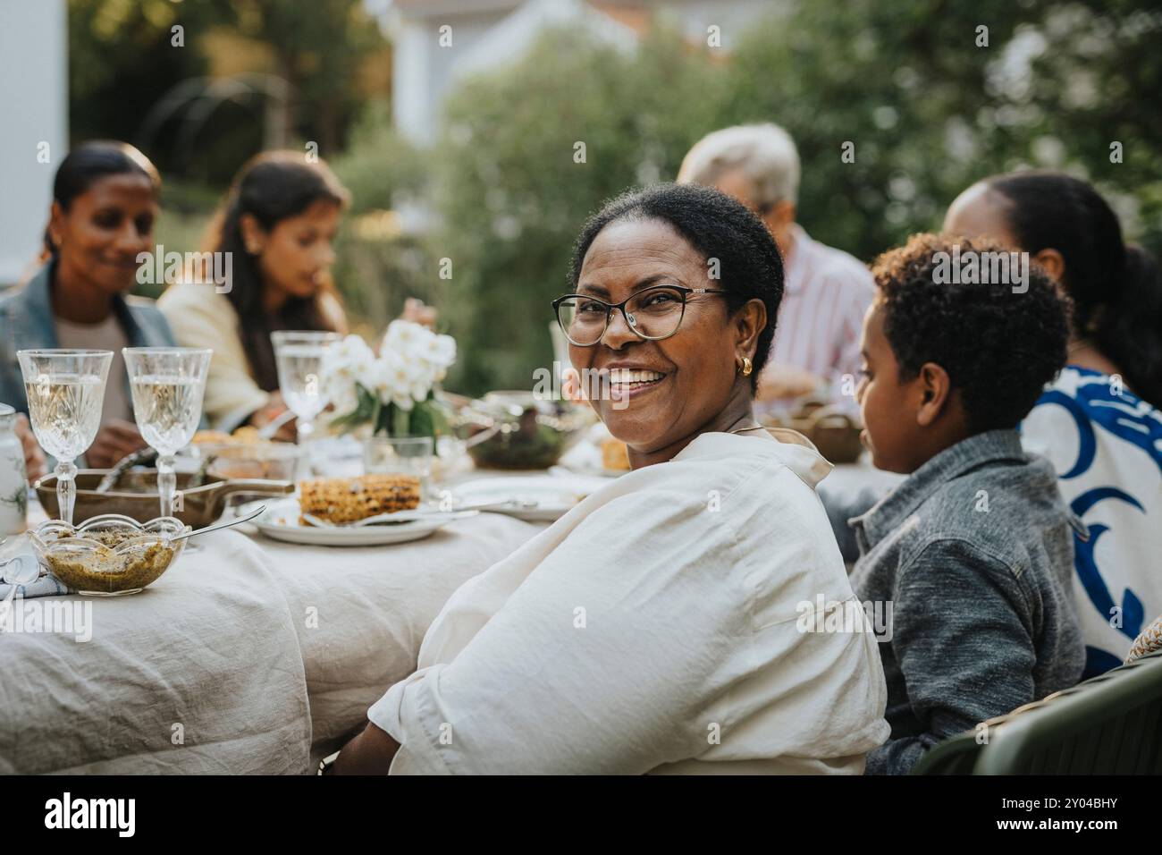 Porträt einer glücklichen Seniorin mit Brille, die über die Schulter schaut, während sie mit Familienmitgliedern beim Mittagessen sitzt Stockfoto