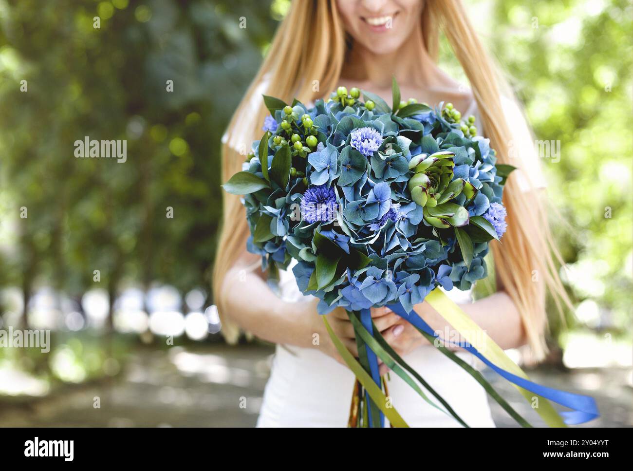 Fruchtige lächelnde junge, langhaarige blonde Frau, die einen schönen eleganten Blumenstrauß mit grünen und blauen Blumen mit Bändern hält, während sie im Park mit GREE steht Stockfoto