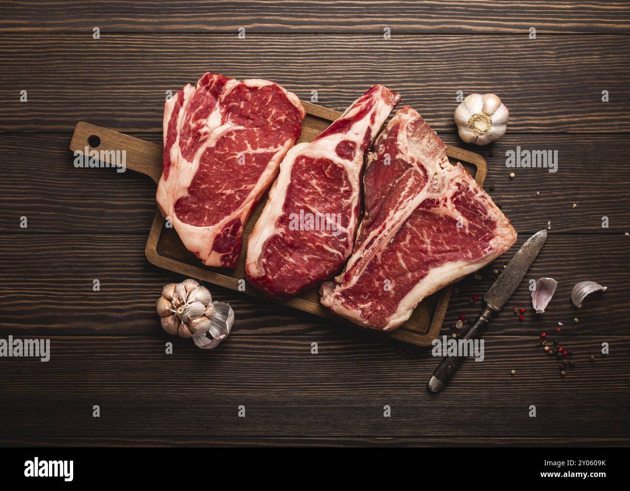 Set aus drei verschiedenen Arten roher Fleischsteaks: Ribeye, T-Bone, Cowboy auf Schneidebrett mit Messer und Gewürzen, Holzhintergrund. Auswahl an gereiften Steaks Stockfoto