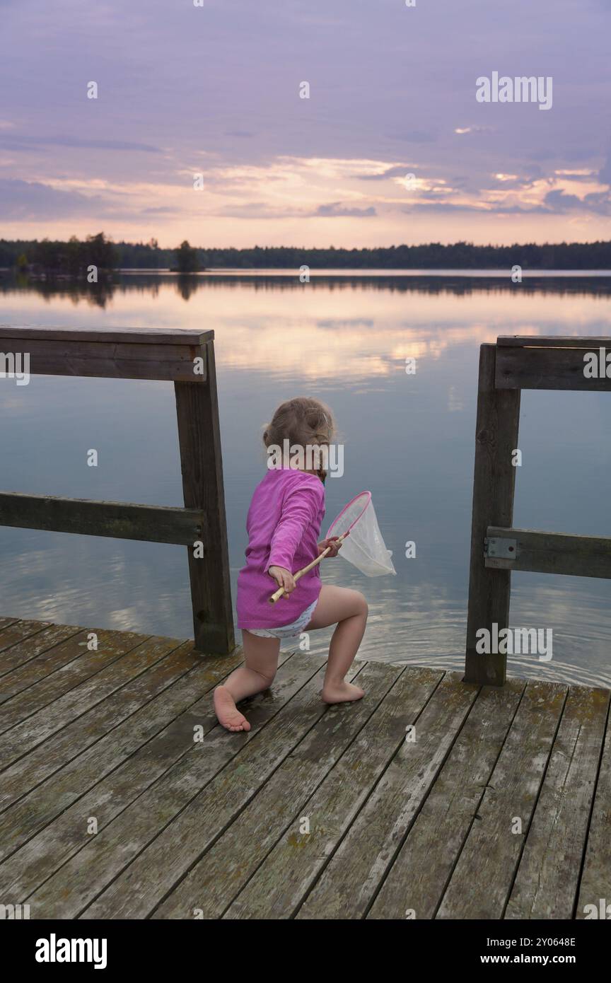 Niedliches kleines Mädchen kniet auf den Holzplanken eines Piers und fischt mit einem Landenetz im Licht eines skandinavischen Sommerabends Stockfoto