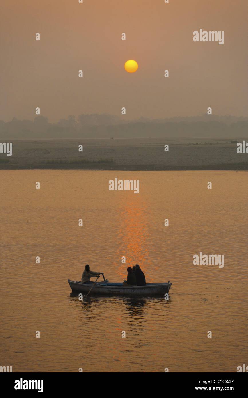 Ein Boot bringt Passagiere auf eine Bootstour bei Sonnenuntergang auf dem heiligen Ganges, während die Sonne von der Wasseroberfläche in Varanasi, Indien, Asien reflektiert wird Stockfoto