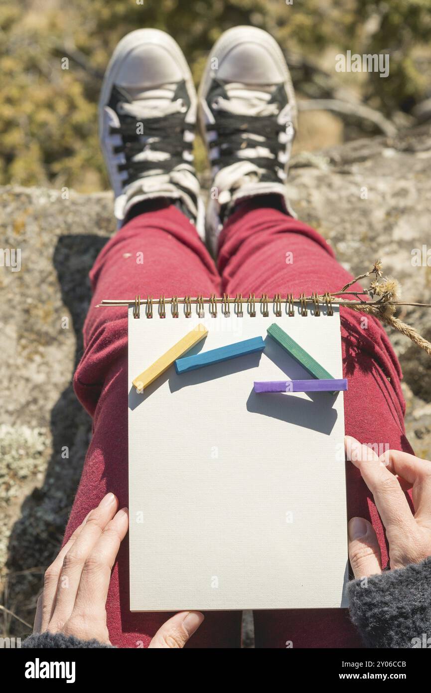 Blick von der ersten Person auf dem weiblichen Schoß ist ein Notizbuch mit Kreiden zum Zeichnen von Pastellfarben. Weibliche Hände, die ein Notizbuch in der Natur halten. Ein Mädchen sitzt Stockfoto