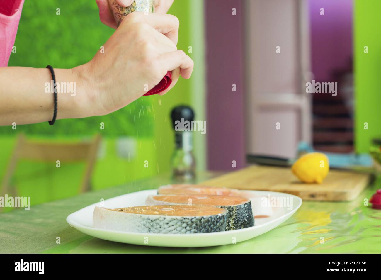 Frauenhände besprühen aus der Handmühle mit Gewürzen zwei rohe Steak vom Lachs, die auf einem weißen Teller in einer Küche in Grüntönen liegen. Das Konzept o Stockfoto