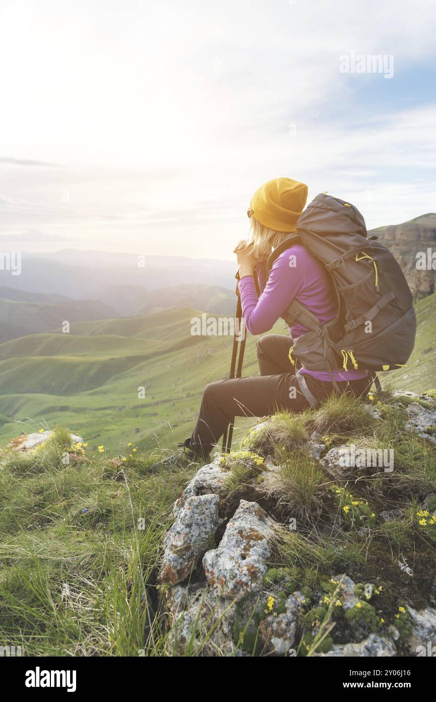 Lächelnde Reiseteilnehmerin mit gelbem Hut und Sonnenbrille, die am Fuße epischer Felsen mit einem Rucksack sitzt und wegschaut Stockfoto