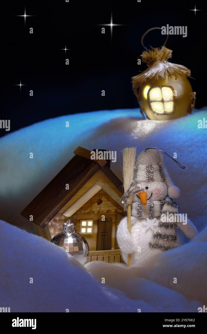 Helles konzeptionelles Weihnachtsstillleben mit einem Schneemann, der neben einem Holzhaus unter einem schneebedeckten Hügel steht. Künstlerische Weihnachtskarte, Rea Stockfoto