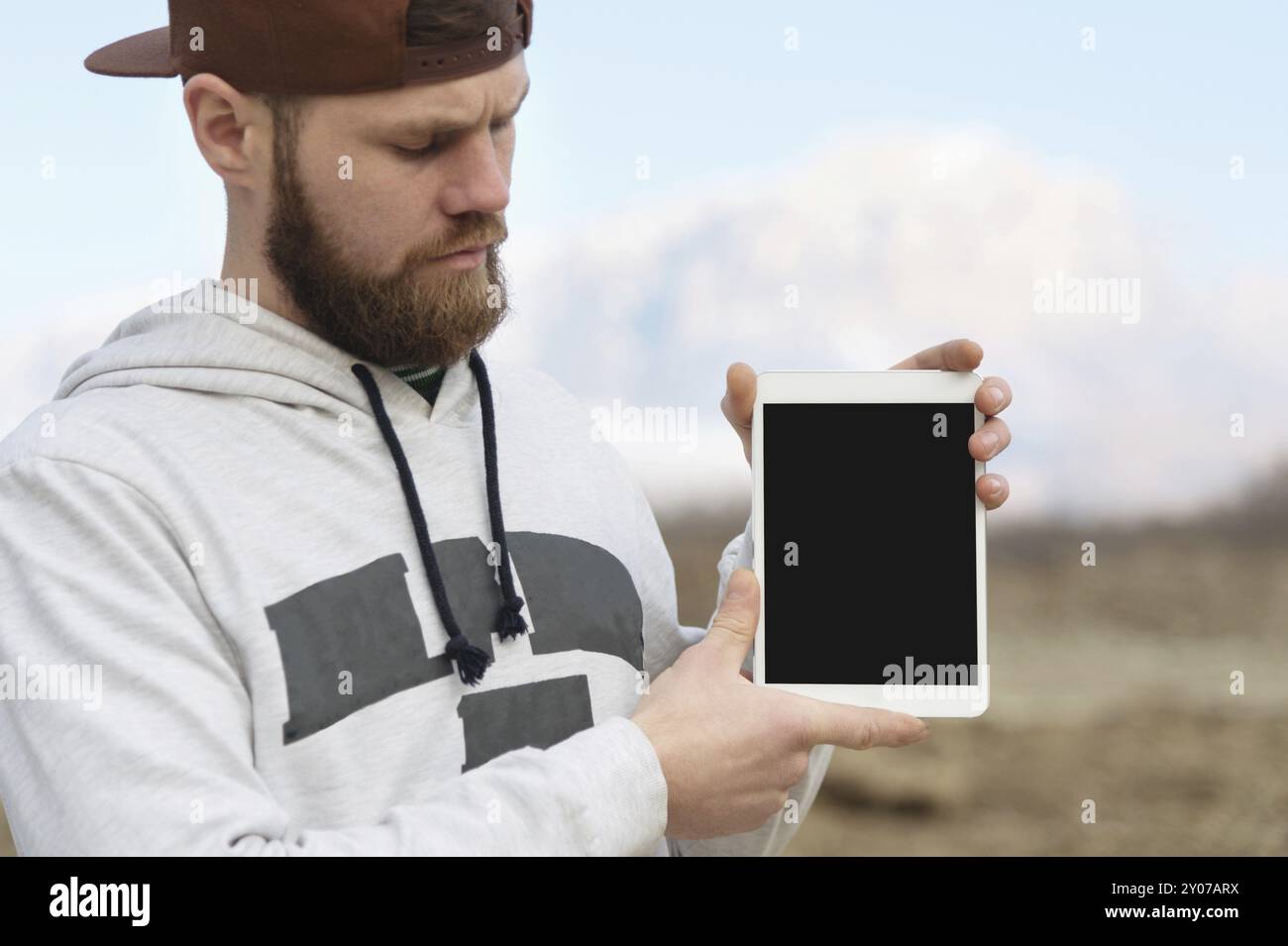 Nahaufnahme Porträt eines Hipsters in brauner Kappe im Freien hält einen weißen Tablet-pc in den Händen. Ein bärtiger Mann schaut sich die Tafel an. Hintergrundunschärfe Stockfoto